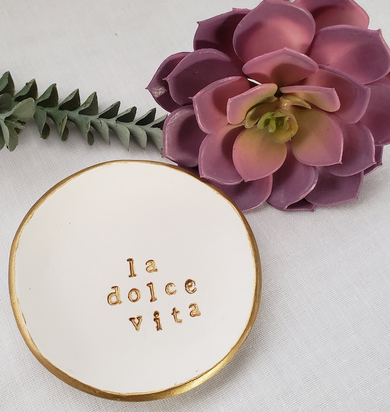 May include: White ceramic ring dish with a gold rim. The dish has the phrase "la dolce vita" written in gold lettering.