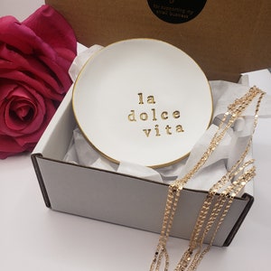 May include: White ceramic trinket dish with a gold rim and the phrase "la dolce vita" written in gold lettering. The dish is in a white box with tissue paper and a gold chain necklace.