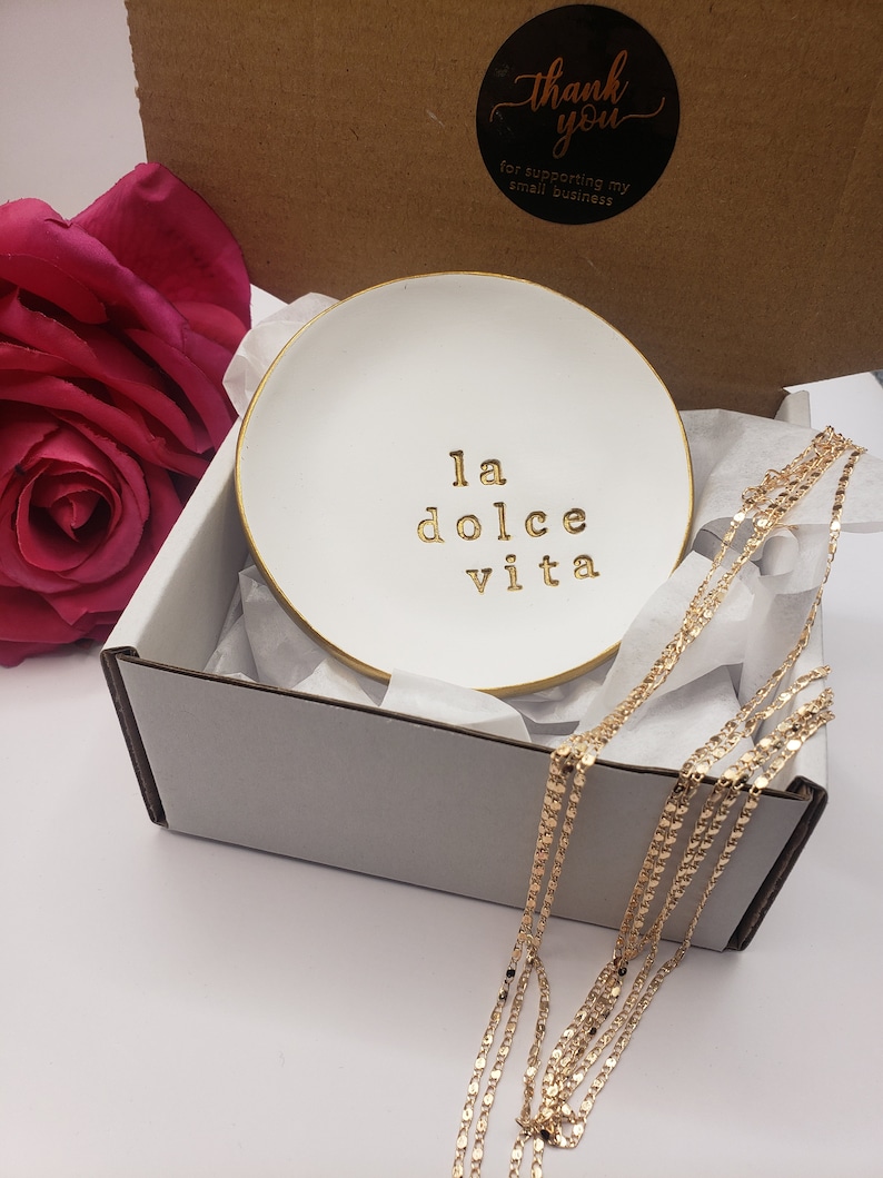 May include: White ceramic trinket dish with a gold rim and the phrase "la dolce vita" written in gold lettering. The dish is in a white box with tissue paper and a gold chain necklace.