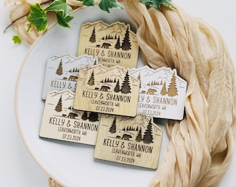 May include: Wooden magnets with a mountain and bear design. Each magnet features the names "Kelly & Shannon", the location "Leavenworth, WA", and the date "07.23.2024". The magnets are arranged on a white plate with a beige cloth and green leaves.