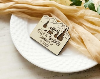 May include: A wooden wedding favor with a mountain and bear design. The favor is engraved with the names "Kelly & Shannon", the location "Leavenworth, WA", and the date "07.23.2024". The favor is on a white plate with a beige cloth background.