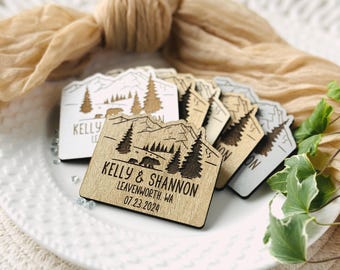 May include: A collection of wooden wedding favors with a mountain and bear design. The favors are engraved with the names "Kelly & Shannon", the location "Leavenworth, WA", and the date "07.23.2024". The favors are in various shades of wood and gray.