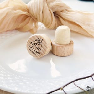 May include: Two wooden bottle stoppers with a light wood finish. One has a natural cork top. The other is engraved with the names "Britney and Hugo" and the date "06.08.27" along with a graphic of two toasting glasses.