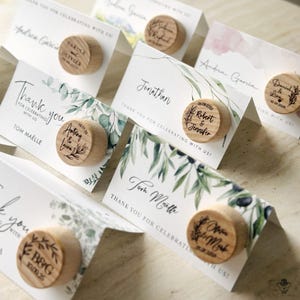 Puede incluir: Tarjetas de lugar blancas con follaje verde y diseños de acuarela. Cada tarjeta tiene un tapón de botella de madera con un mensaje personalizado y una fecha. El texto en los tapones incluye nombres y fechas, como "Robert & Jennifer 08.08.2023".