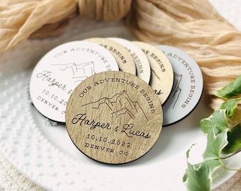 May include: A collection of round wooden coasters with a mountain design and the text "Our Adventure Begins." The coasters are personalized with names, a date, and location. The coasters are in various shades of wood and gray.
