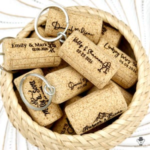 May include: A selection of personalised cork keyrings with silver keyrings, presented in a woven basket. Each cork is engraved with names and dates, some also feature a wine glass or mountain graphic. The keyrings are a light brown colour.