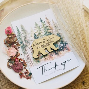 Pode incluir: Um cartão de agradecimento de casamento com um recorte de urso de madeira. O cartão apresenta uma cena de floresta em aquarela e as palavras "Thank you". O recorte de urso tem os nomes "Nicole & Ryan", a data "08.10.2024" e a localização "Dolores, Colorado".
