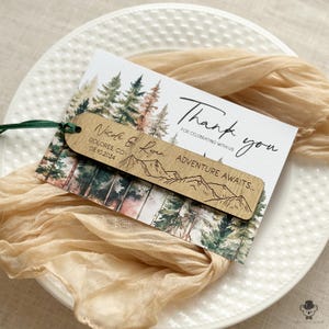 Könnte beinhalten: Eine Hochzeitsdankeskarte mit einer Waldszene und den Worten "Danke" und "Abenteuer erwartet...". Ein Holzlesezeichen mit einem Bergketten-Design ist befestigt. Die Karte enthält die Namen Nicole & Ryan und das Datum 08.10.2024.