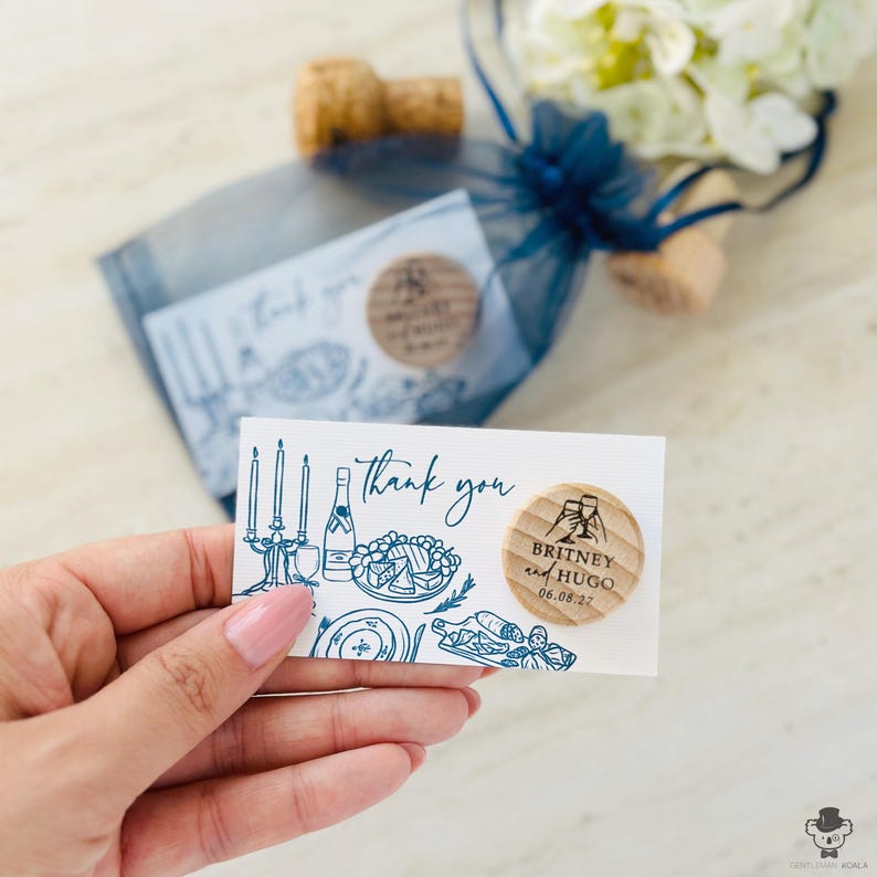 A hand holding a small card with a message and a cork, along with a blue ribbon and a bouquet of white flowers.