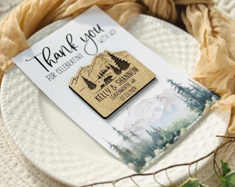 May include: A wedding thank you card with a wooden magnet. The card reads "Thank you for celebrating with us!" The magnet features a mountain and tree design with the names "Kelly & Shannon" and the date "07.23.2024".
