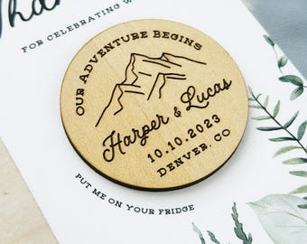 May include: A round wooden magnet with engraved text and mountain graphic. The text reads "Our Adventure Begins," the names "Harper & Lucas," the date "10.10.2023," and "Denver, CO." The magnet is on a white card with the text "Thank you" and "Put me on your fridge."