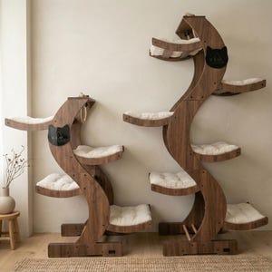 May include: Two wooden cat trees with multiple platforms and plush, cream-colored cushions. The trees have a curved, organic design and feature a black cat silhouette with the letters "W.E.O." on a plaque. A rope and a small toy are attached to the top.