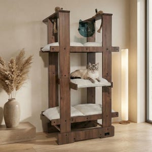 May include: A dark wooden cat tree with three platforms, each with a white cushion. A cat is resting on the middle level. The design incorporates rope detailing and a logo. This cat furniture offers a comfortable space for cats to relax and play.