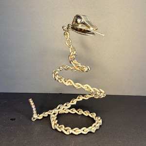 Op de afbeelding: Een handgemaakte metalen sculptuur van een slang. Het lichaam van de slang is gevormd uit een ketting, geschilderd in een verweerde witte kleur. De kop is zilverkleurig met een gedetailleerd ontwerp. De sculptuur wordt gepresenteerd tegen een neutrale achtergrond.