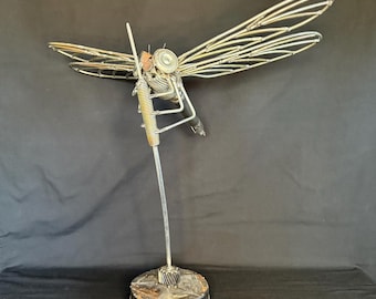 Pode incluir: Escultura de libélula de metal com asas e corpo de arame detalhados. A escultura é montada sobre uma base redonda em forma de engrenagem. O corpo da libélula apresenta uma mola e outros componentes mecânicos.