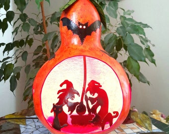 May include: A gourd lamp with a Halloween theme. The gourd is painted orange and features a silhouette of a bat, two witches stirring a cauldron, and a black cat. The lamp is lit from within, casting a warm glow.