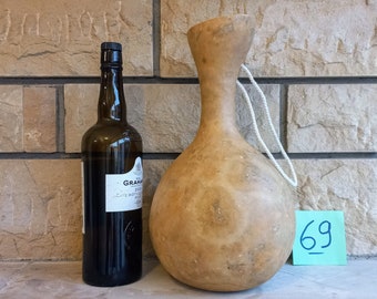 Könnte beinhalten: Ein hellbrauner Kürbis mit einem weißen Seilgriff, neben einer dunkelbraunen Glasflasche mit einem Etikett, auf dem "Graham's 20 Year Old Tawny Port, Late Bottled Vintage" steht. Der Kürbis und die Flasche stehen auf einer hellbraunen Ziegelstein-Oberfläche. Ein blaues Schild mit der Nummer "69" befindet sich auf der rechten Seite des Bildes.