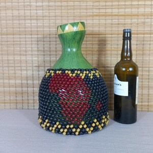 May include: A decorative vase with a green neck and a beaded body featuring a red African continent design. The vase is next to a brown glass bottle with a label.