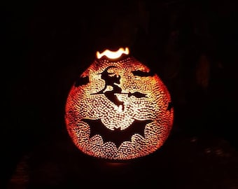 May include: A carved gourd lamp with a Halloween theme. The gourd is lit from within, casting a warm glow through the intricate carvings of bats and a witch flying on a broomstick.