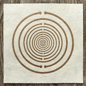 Op de afbeelding: Een vierkant stuk bruin papier met een concentrisch cirkelontwerp in bruin. De cirkels zijn gelijkmatig verdeeld en creëren een cirkelvormig patroon.