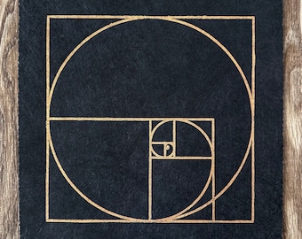 Può includere: Opera d'arte quadrata nera con un design a spirale dorata, una rappresentazione visiva della sequenza di Fibonacci. L'opera d'arte ha una superficie strutturata ed è ambientata su uno sfondo di legno. Le linee dorate creano un motivo geometrico.