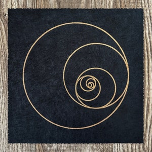Könnte beinhalten: Ein schwarzer quadratischer Untersetzer mit einem goldenen Spiraldesign. Die Spirale besteht aus konzentrischen Kreisen, die immer kleiner werden, je näher sie dem Zentrum kommen.
