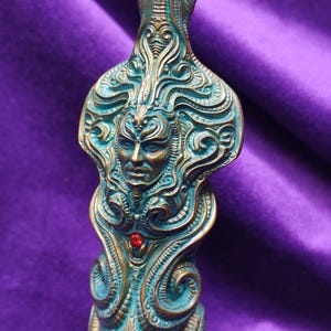May include: Ornate bronze-toned sculpture with teal accents, featuring a detailed face and swirling patterns. A small red gem is set below the face. The sculpture is set against a purple background.