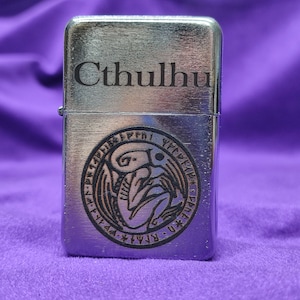May include: A silver lighter with a black engraved design of Cthulhu and a circle of runes. The lighter has the word "Cthulhu" engraved on the top.