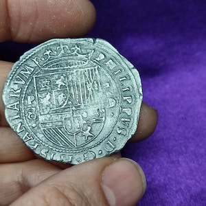 Pode incluir: Uma moeda de prata com um brasão de armas e a inscrição "REX PHILIP. II. HISPANIARUM", que se traduz como "Rei Filipe II de Espanha".