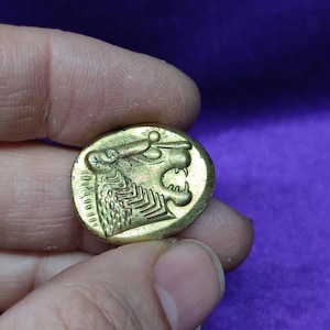 May include: A gold-colored coin with a detailed image of a lion's head facing right. The lion has a crown on its head and a mane of hair. The coin is in good condition and has a smooth surface.