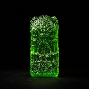May include: A translucent, vibrant green rectangular object featuring a detailed, relief-style sculpted face with tentacle-like features and intricate, swirling patterns covering the entire surface, set against a solid black background.