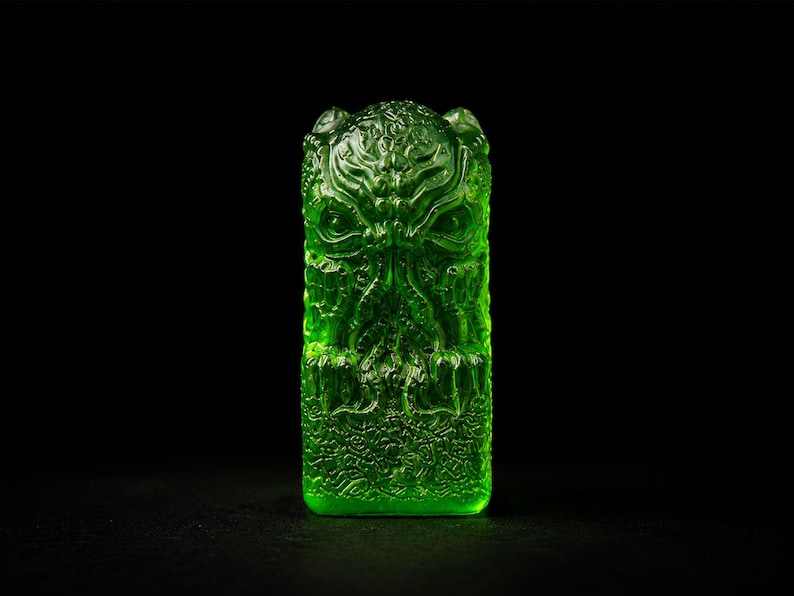 May include: A translucent, vibrant green rectangular object featuring a detailed, relief-style sculpted face with tentacle-like features and intricate, swirling patterns covering the entire surface, set against a solid black background.
