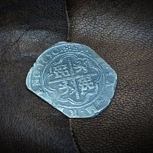 Puede incluir: Una moneda de plata con una cruz y un diseño floral. La moneda está sobre una superficie de cuero marrón.