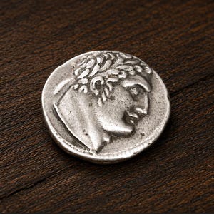 May include: A circular silver-coloured coin featuring a relief profile of a person with curly hair facing right. The coin has a weathered, metallic texture with raised details on the surface, showcasing a classic design reminiscent of ancient currency.