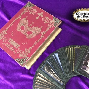 May include: A red tarot deck box with a gold ornate design and a black mask. The box is open and the tarot cards are fanned out on a purple surface. The cards are black with gold accents and feature a dark, gothic style.