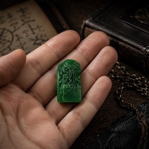 May include: A small, rectangular, emerald green figurine featuring a detailed, relief-carved creature with tentacles extending from its face. The object has a smooth, polished surface and is shown resting in the center of an open palm.