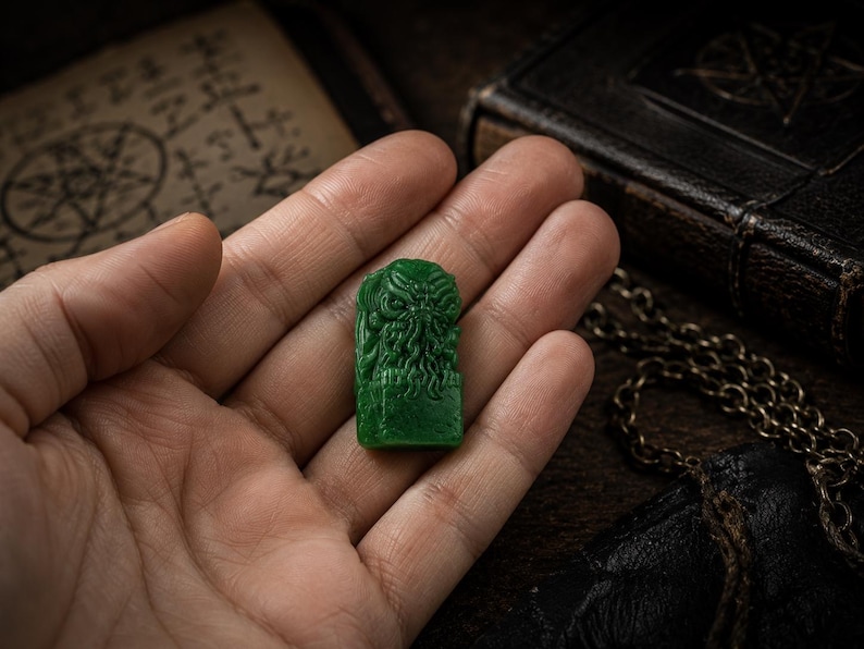 May include: A small, rectangular, emerald green figurine featuring a detailed, relief-carved creature with tentacles extending from its face. The object has a smooth, polished surface and is shown resting in the center of an open palm.