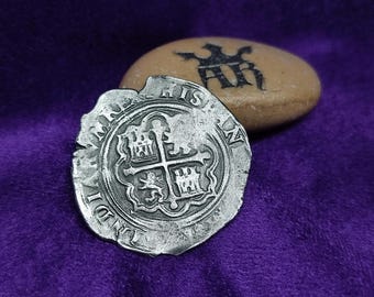 May include: A silver coin with the inscription "REX HISPANIARUM" and a coat of arms featuring a cross and four castles. The coin is slightly worn and has a rough texture.