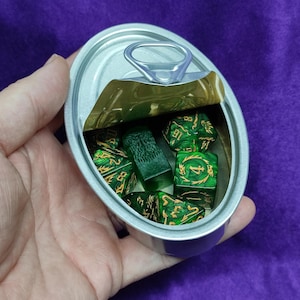 Puede incluir: Una lata plateada con una anilla, llena de dados verdes y dorados. Los dados son de varias formas y tamaños, con números dorados. La lata se sostiene en una mano sobre un fondo morado. Los dados son para juegos de mesa.
