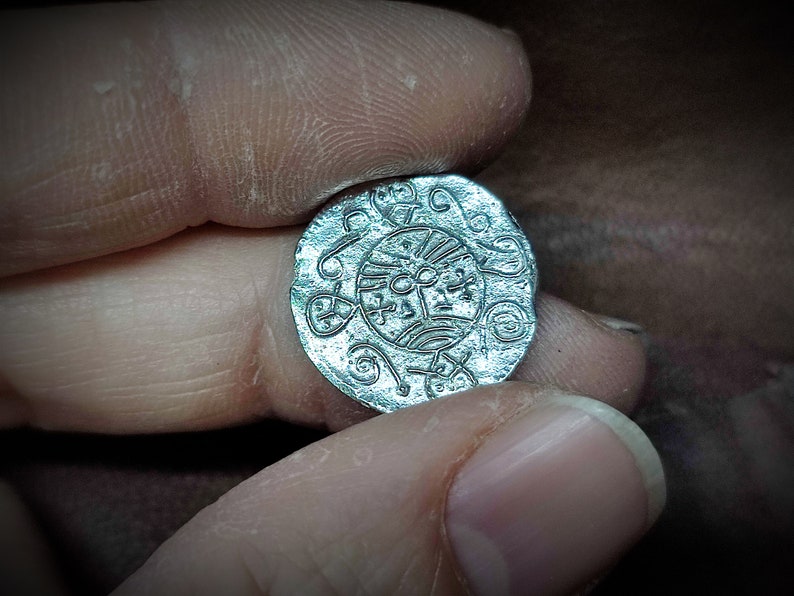 Może przedstawiać: Srebrna moneta z wytłoczonym wzorem przedstawiającym centralny motyw przypominający słońce otoczony wirującymi wzorami. Moneta ma lekko nieregularny, okrągły kształt i wydaje się być antykiem lub repliką.