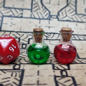 Könnte beinhalten: Zwei kleine Glasflaschen mit Korkstopfen, eine mit grüner Flüssigkeit gefüllt und die andere mit roter Flüssigkeit. Ein roter zwanzigseitiger Würfel befindet sich im Vordergrund.