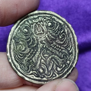 Könnte beinhalten: Eine runde, silberfarbene Münze mit einem komplizierten, geprägten Design aus wirbelnden Mustern und Formen. Das Design scheint eine stilisierte Darstellung einer Kreatur oder eines mythischen Wesens zu sein.