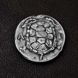 Op de afbeelding: Een ronde, zilverkleurige munt met in het midden een reliëf van een schildpadschild. De letters A en I staan in reliëf aan de linker- en rechterkant van het schild. De munt heeft een verweerd, getextureerd oppervlak met een metalen afwerking.