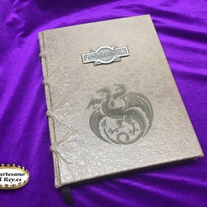 Può includere: Un diario rilegato in pelle marrone con un drago argentato in rilievo sulla copertina. Il diario ha una chiusura in metallo argentato con il testo "The Dragon's Den" su di essa.