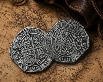 May include: Two silver-colored metal coins with embossed heraldic shield designs and Latin inscriptions, resting near a dark brown leather drawstring pouch and a portion of a brass compass on a vintage-style map with nautical illustrations.