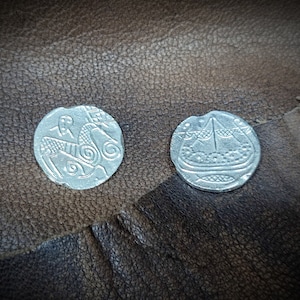 Może przedstawiać: Dwie srebrne monety ze skomplikowanymi wzorami. Jedna moneta przedstawia konia i jeźdźca, druga ł&oacute;dź ze szczeg&oacute;łowym wzorem. Monety są prezentowane na teksturowanej, brązowej sk&oacute;rzanej powierzchni, sugerując motyw antyczny lub historyczny.