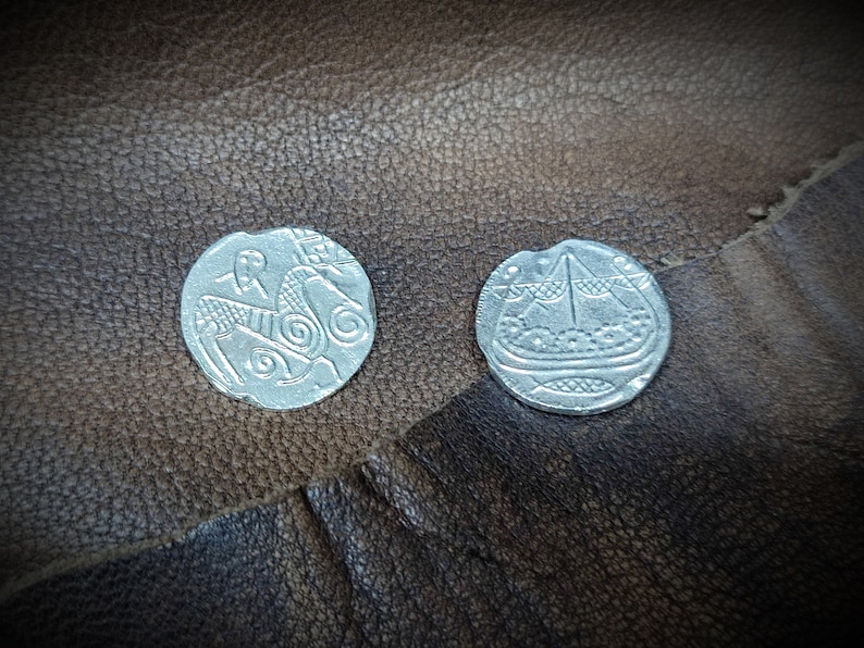 Może przedstawiać: Dwie srebrne monety ze skomplikowanymi wzorami. Jedna moneta przedstawia konia i jeźdźca, druga ł&oacute;dź ze szczeg&oacute;łowym wzorem. Monety są prezentowane na teksturowanej, brązowej sk&oacute;rzanej powierzchni, sugerując motyw antyczny lub historyczny.