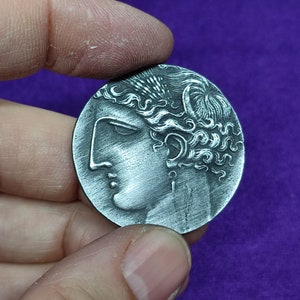 Puede incluir: Una moneda plateada con un perfil detallado de una mujer con el pelo rizado. La moneda tiene un acabado liso y metálico.