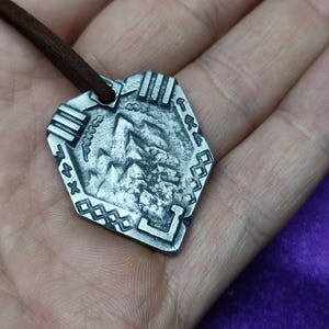 Puede incluir: Un collar con un colgante de plata con un diseño detallado de una cordillera y un borde geométrico. El colgante está unido a un cordón de cuero marrón.