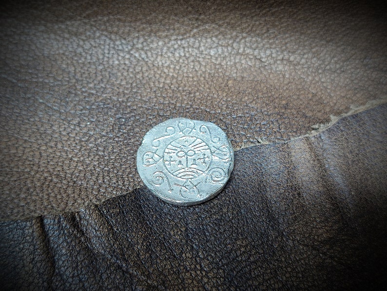 Może przedstawiać: Srebrna moneta ze skomplikowanym wzorem przedstawiającym centralny symbol otoczony wirującymi wzorami. Moneta jest umieszczona na teksturowanej, brązowej sk&oacute;rzanej powierzchni, tworząc kontrast między metalicznym połyskiem a rustykalnym tłem.